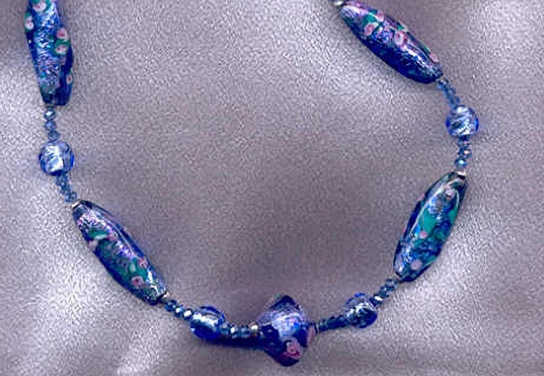 May include: A blue and white glass bead necklace with floral patterns. The necklace is made of small, round beads and larger, oval beads.