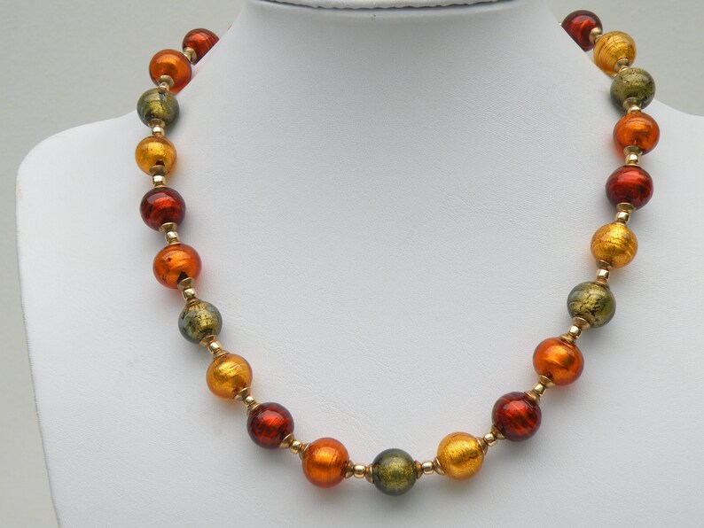 May include: A necklace made of round, multi-colored glass beads. The beads are shades of green, orange, and red. The beads are strung on a gold chain.
