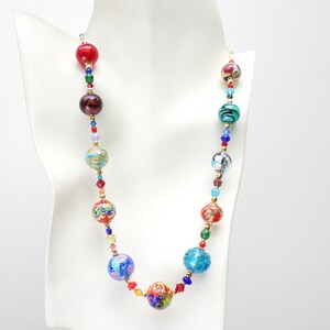 May include: A vibrant beaded necklace featuring large, patterned beads in red, blue, turquoise, and gold. The necklace includes smaller, colourful beads and gold-toned spacers. The necklace is displayed on a white mannequin.