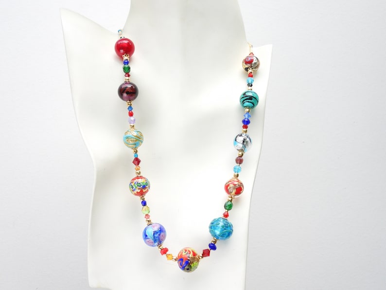 May include: A vibrant beaded necklace featuring large, patterned beads in red, blue, turquoise, and gold. The necklace includes smaller, colourful beads and gold-toned spacers. The necklace is displayed on a white mannequin.