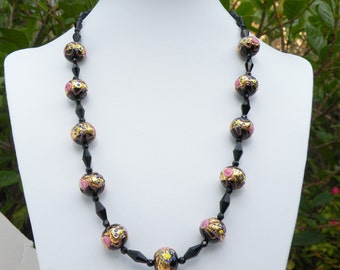 May include: A black and gold necklace with pink and blue accents. The necklace is made of round beads with intricate designs.