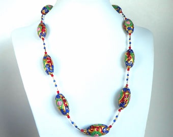 Puede incluir: Un collar largo con grandes cuentas ovaladas en oro, azul, rojo y verde. Las cuentas están ensartadas con cuentas más pequeñas rojas, azules y doradas. El collar se exhibe en un maniquí blanco.