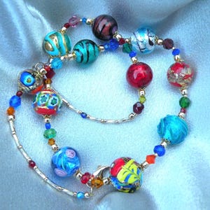 May include: A vibrant beaded necklace featuring glass beads in blue, red, gold, and green. The beads, with intricate designs, are strung on a silver chain. The necklace is displayed on a light blue satin fabric. The necklace is a statement piece.