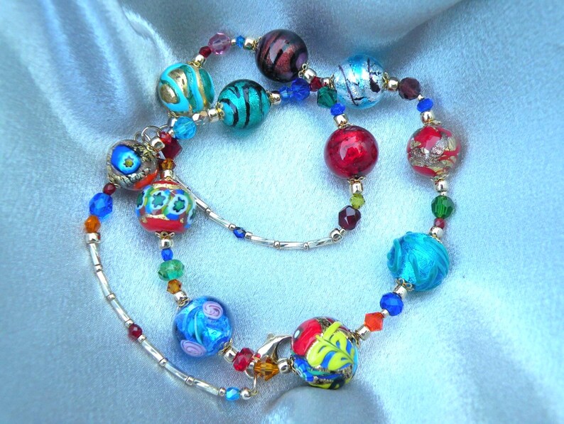May include: A vibrant beaded necklace featuring glass beads in blue, red, gold, and green. The beads, with intricate designs, are strung on a silver chain. The necklace is displayed on a light blue satin fabric. The necklace is a statement piece.