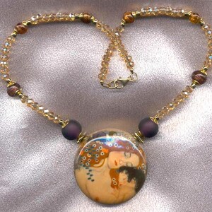 Puede incluir: Un collar dorado con un colgante redondo que representa una imagen art&iacute;stica de dos figuras abraz&aacute;ndose. El collar tiene una cadena de cuentas con cristales color &aacute;mbar y cuentas moradas oscuras. El cierre es dorado.