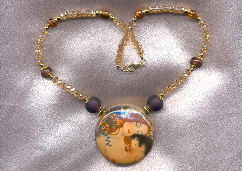 Puede incluir: Un collar dorado con un colgante redondo que representa una imagen art&iacute;stica de dos figuras abraz&aacute;ndose. El collar tiene una cadena de cuentas con cristales color &aacute;mbar y cuentas moradas oscuras. El cierre es dorado.