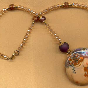 Puede incluir: Un collar dorado con un colgante grande y redondo que representa una pintura de dos figuras abraz&aacute;ndose. El collar tiene una cadena de cuentas con peque&ntilde;os cristales transparentes y cuentas rojas oscuras. El cierre es dorado.