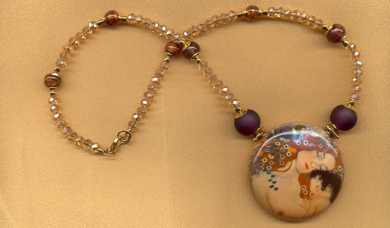 Puede incluir: Un collar dorado con un colgante grande y redondo que representa una pintura de dos figuras abraz&aacute;ndose. El collar tiene una cadena de cuentas con peque&ntilde;os cristales transparentes y cuentas rojas oscuras. El cierre es dorado.
