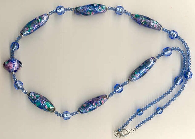 May include: A blue and purple glass bead necklace with a silver clasp. The necklace features a variety of blue and purple beads, including some with floral patterns.