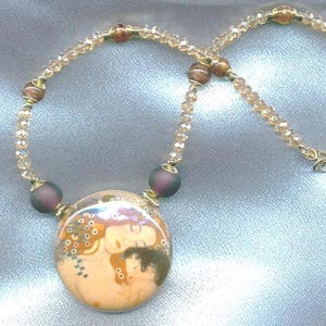 Puede incluir: Un collar dorado con un colgante que representa una imagen art&iacute;stica de dos figuras. El collar tiene una cadena de cuentas con peque&ntilde;as cuentas transparentes y cuentas m&aacute;s grandes de color p&uacute;rpura oscuro. El colgante es redondo con un fondo color crema.