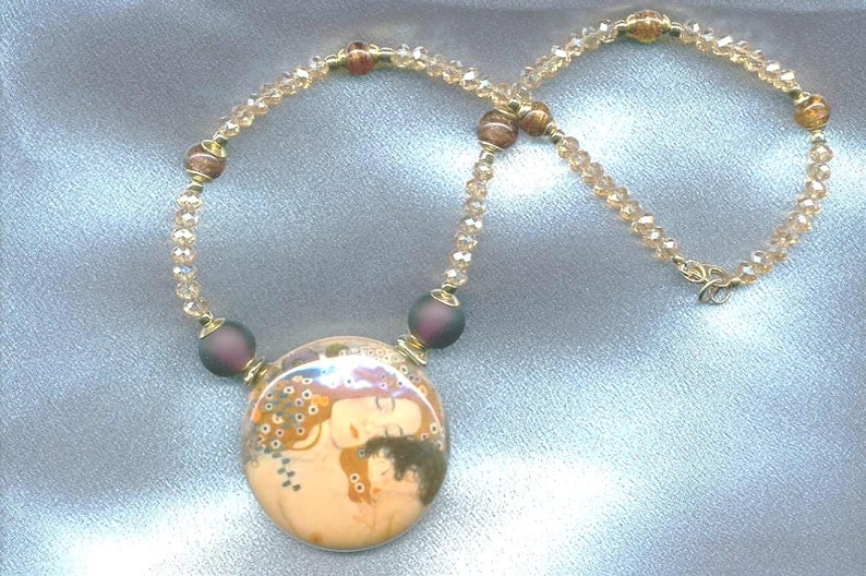 Puede incluir: Un collar dorado con un colgante que representa una imagen art&iacute;stica de dos figuras. El collar tiene una cadena de cuentas con peque&ntilde;as cuentas transparentes y cuentas m&aacute;s grandes de color p&uacute;rpura oscuro. El colgante es redondo con un fondo color crema.