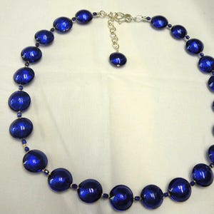 Peut inclure: Un collier compos&eacute; de grosses perles rondes bleu fonc&eacute;. Les perles sont enfil&eacute;es avec de plus petites perles dor&eacute;es et un fermoir &agrave; cha&icirc;ne argent&eacute;. Le collier est pr&eacute;sent&eacute; sur une surface blanche.