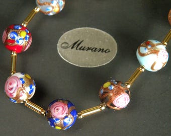 May include: A necklace featuring colorful Murano glass beads. The beads are round with intricate designs in red, blue, pink, and gold. The beads are strung together with gold-colored metal bars. The word "Murano" is on a gray oval tag.