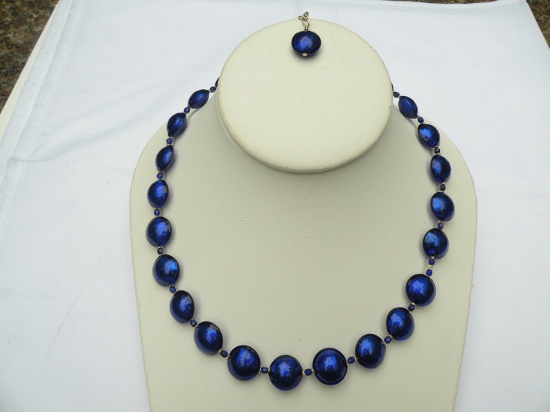 Peut inclure: Un collier de perles bleu fonc&eacute; et un pendentif assorti. Le collier comporte de grosses perles rondes entrecoup&eacute;es de perles ovales plus petites. Le pendentif est une seule perle ronde. Les bijoux sont expos&eacute;s sur un buste blanc.