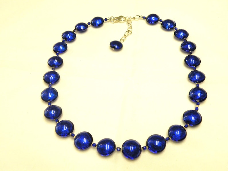 Peut inclure: Un collier compos&eacute; de grandes perles rondes en verre bleu fonc&eacute;. Les perles sont enfil&eacute;es avec de petites perles dor&eacute;es et un fermoir argent&eacute;. Le collier est pr&eacute;sent&eacute; sur un fond jaune clair.