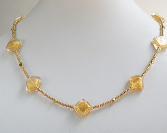 May include: A gold necklace with square-shaped glass beads that have gold leaf accents. The necklace is on a white background.