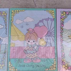 May include: Three framed coloring pages featuring Precious Moments characters. The first page shows a girl with blonde hair holding a white goose. The second page shows a girl dressed as a clown holding a pink heart-shaped balloon. The third page shows a girl with blonde hair wearing a crown and a pink dress standing in front of a pink castle. The text on the second page reads "Smile Along The Way."