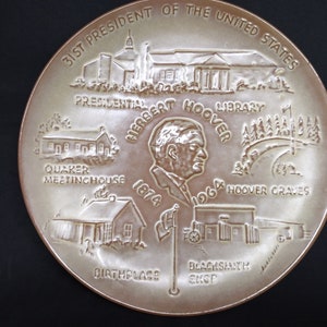 May include: A white ceramic plate with a brown border depicting the life of Herbert Hoover, the 31st President of the United States. The plate features images of his birthplace, Quaker Meetinghouse, Presidential Library, and Hoover Graves. The text "Herbert Hoover 1874 1964" is inscribed on the plate.