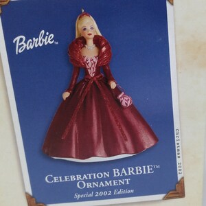 May include: A red and white Barbie doll ornament with a red dress and a white fur collar. The ornament is labeled "Celebration Barbie Ornament Special 2002 Edition".