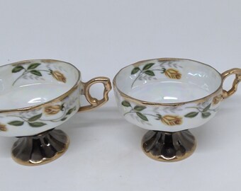 May include: Two white porcelain teacups with gold trim and yellow rose floral designs. The cups have a pedestal base and a gold handle.