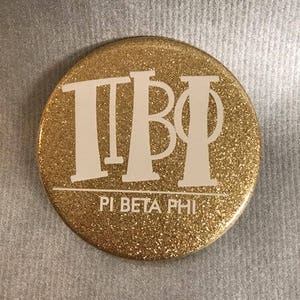 May include: Gold glitter button with white letters that say "ΠΒΦ" and "Pi Beta Phi".