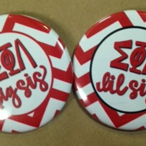 May include: Two round buttons with a red and white chevron pattern. One button reads "ΣΦΛ big sis" in red script, and the other reads "ΣΦΛ lil sis". The buttons have a white center with black borders.