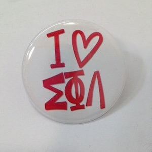 May include: A white button with red text that reads "I ♥ ΣΦΛ". The button has a glossy finish and a simple, clean design. The red text is in a bold, sans-serif font.