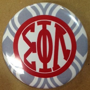 May include: A round button with a white and gray chevron pattern background. The center features a red circle with white Greek letters. The button is a pin-back style.