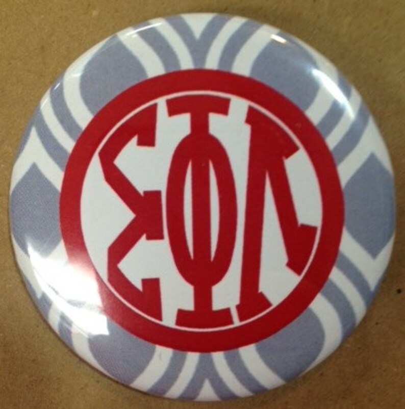 May include: A round button with a white and gray chevron pattern background. The center features a red circle with white Greek letters. The button is a pin-back style.