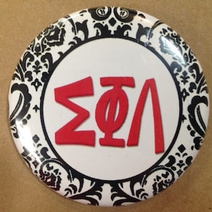 May include: A white button with a black and white damask pattern border. The center of the button has a red Greek letter 'ΣΦΛ' on a white background.
