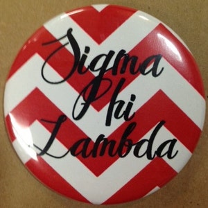 May include: A round button with a red and white chevron pattern. The button features the words "Sigma Phi Lambda" in black script across the center. The button is a decorative item.