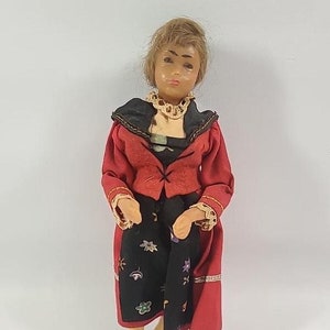 May include: A vintage doll wearing a red jacket, black skirt with floral embroidery, and black shoes. The doll has brown hair and a painted face.