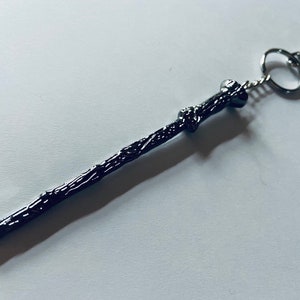 May include: A dark, metallic, wand-shaped keychain featuring a textured, branch-like surface with a gnarled handle design. The wand is attached to a silver-toned metal split ring and a small lobster claw clasp, providing a functional attachment point.