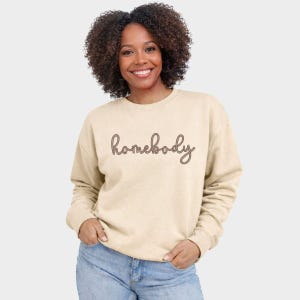 May include: A beige sweatshirt with the word "homebody" in a cursive font. The long-sleeved crew neck shirt is worn by a person with curly dark hair and blue jeans. The person is smiling and looking at the camera.