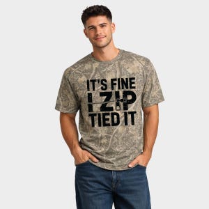 May include: A camouflage-patterned t-shirt with the text "IT'S FINE I ZIP TIED IT" in black. The shirt is short-sleeved and has a crew neck. The overall color scheme is shades of green and brown.