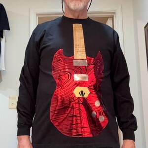 May include: Black long-sleeve shirt featuring a red guitar graphic. The guitar design has a swirling pattern and a wooden neck. The shirt is made of a soft material.