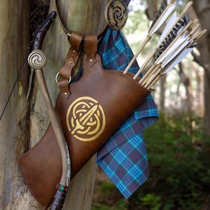 May include: A brown leather quiver with a gold Celtic design, holding several arrows with white and black fletching. A matching bow with a Celtic design is also present, along with a blue and green plaid fabric.