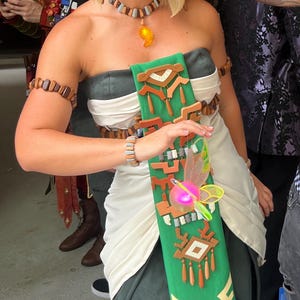 Pode incluir: Uma mulher em um traje de cosplay, com um vestido verde e branco com detalhes em madeira. Ela usa uma tiara, colar e pulseiras de madeira. Uma faixa verde decorativa com desenhos geométricos e uma fada brilhante completa o visual.