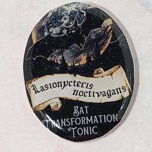 May include: An oval-shaped button with a black background featuring a bat illustration and the text "Lasionycteris noctivagans BAT TRANSFORMATION TONIC" in a vintage font. The button has a glossy finish.