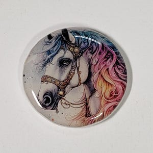 May include: A round, glossy button featuring a detailed illustration of a horse's head. The horse has a white face, adorned with a golden bridle. Its mane and forelock are a blend of blue, pink, and yellow hues, creating a colorful effect.