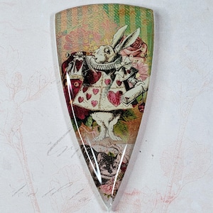May include: A decorative, shield-shaped pendant featuring a vintage illustration of the White Rabbit from Alice in Wonderland. The rabbit is dressed in a red coat with heart details, holding a trumpet and scroll. The background has a floral and striped design.