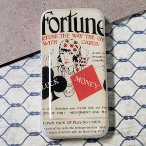 Può includere: Un magnete rettangolare in stile vintage con la parola "Fortune" in nero. L'immagine presenta un'illustrazione di una donna, simboli di carte da gioco e le parole "Luck" e "Money".