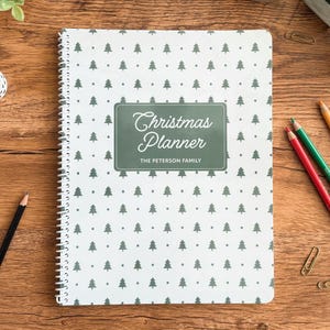 Personalized Christmas Planner with green Christmas tree cover and white spiral binding on a festive desk flat lay. Custom holiday planner for families — track gifts, budget, meals, and to-do lists. Shown with stationery supplies, color pencils, washi tape, and greenery for cozy Christmas aesthetic