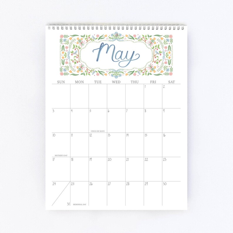 Pu&ograve; includere: Un calendario a spirale per il mese di maggio. La parte superiore presenta la parola &laquo;May&raquo; in corsivo blu, incorniciata da un bordo floreale con accenti verdi, gialli e rosa. La griglia del calendario &egrave; bianca con numeri neri.