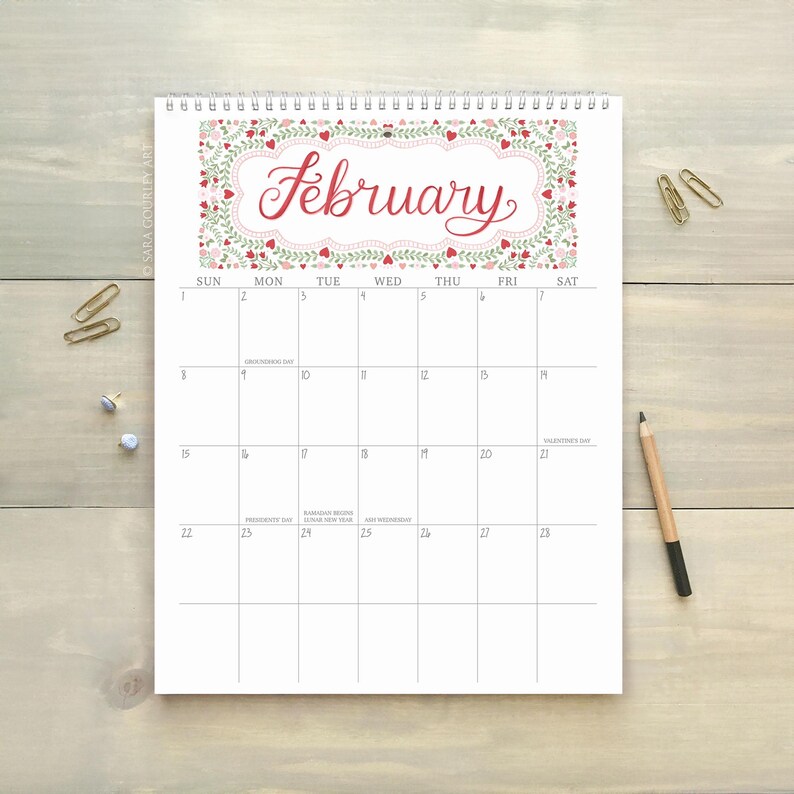 Pu&ograve; includere: Calendario di febbraio con bordo floreale e nome del mese in corsivo. Il calendario &egrave; rilegato a spirale e si trova su una superficie in legno chiaro, con graffette e una matita. Misure in cm.