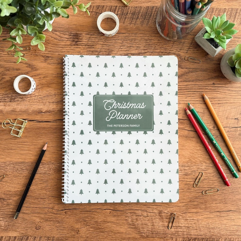Personalized Christmas Planner with green Christmas tree cover and white spiral binding on a festive desk flat lay. Custom holiday planner for families — track gifts, budget, meals, and to-do lists. Shown with stationery supplies, color pencils, washi tape, and greenery for cozy Christmas aesthetic
