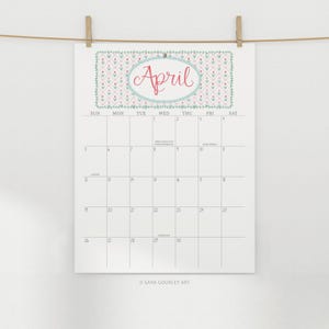 Pu&ograve; includere: Un calendario di aprile bianco con un motivo floreale e il nome del mese in corsivo rosso. Il calendario &egrave; appeso a un filo con mollette di legno. La griglia del calendario &egrave; visibile.