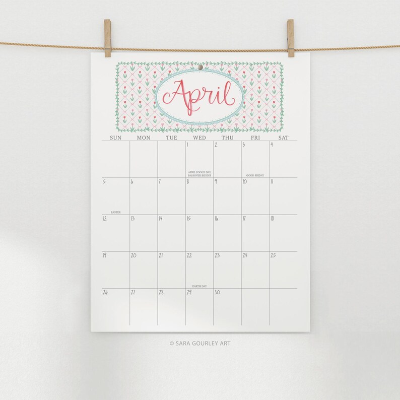 Pu&ograve; includere: Un calendario di aprile bianco con un motivo floreale e il nome del mese in corsivo rosso. Il calendario &egrave; appeso a un filo con mollette di legno. La griglia del calendario &egrave; visibile.