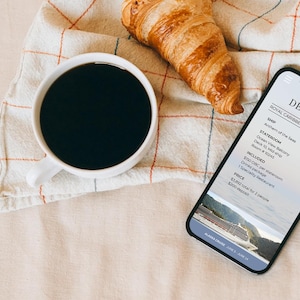 May include: A white coffee cup filled with black coffee, a golden croissant, and a smartphone displaying cruise details. The phone shows information about a Royal Caribbean cruise, including the ship, stateroom, and price.