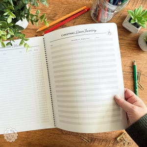 Open personalized Holiday Planner Notebook flat lay showing “Christmas Decor Inventory” and “Christmas Movies” pages. Hand ready to flip the page. Christmas Planner includes sections to track decorations, ornaments, lights, and favorite holiday films. Christmas organization notebook for planning festive décor, movie nights, and seasonal memories — styled on a cozy desk with stationery items.
